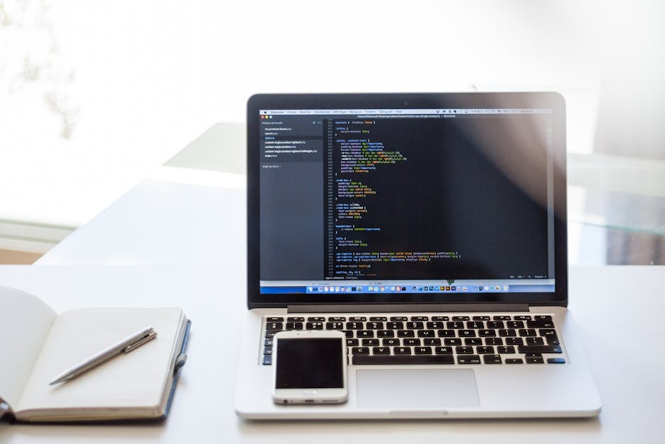 A modern workspace featuring a silver MacBook laptop with an active screen displaying lines of code in a dark editor, positioned on a white desk. To the left of the laptop is an open notebook with a p